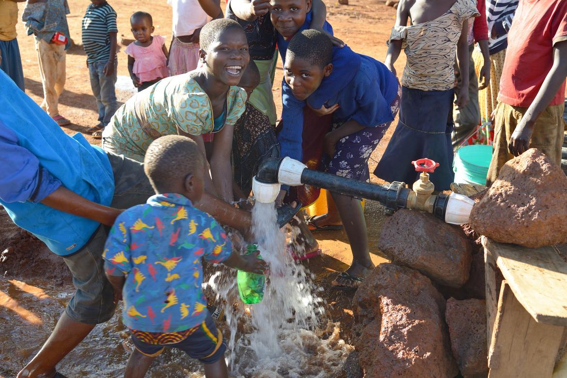 Children stand smiling and screeching at a jet of water coming out of the pipe
