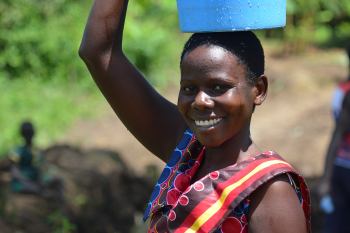 An African woman in a colourful dress carries a blue bucket on her head.